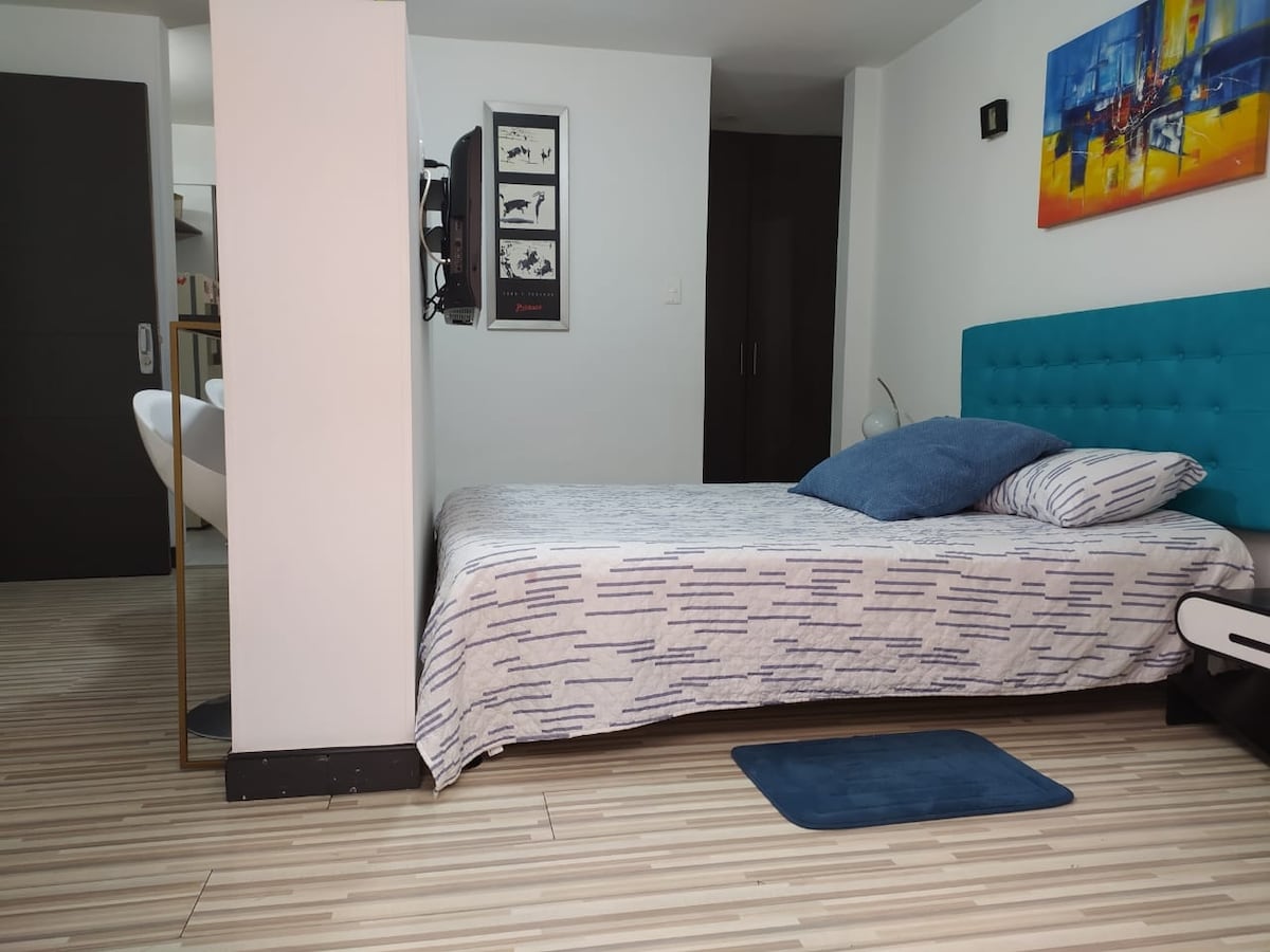 A bright sleeping area features a bed adorned with a patterned blanket and a large cushion. A television is mounted on the wall, and a small rug lies on the floor. The space is complemented by neutral-colored walls and flooring, contributing to the overall calm environment.