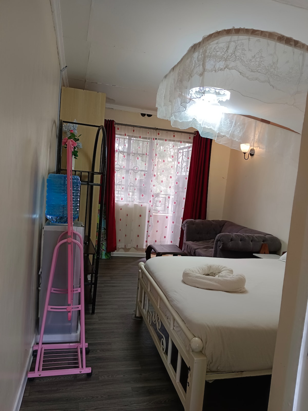 A cozy room is featured, showcasing a spacious bed with a neatly arranged towel. A window with sheer curtains allows natural light to brighten the space, while decorative red drapes frame the window. A small seating area is present with a plush sofa, and a water dispenser is positioned nearby.