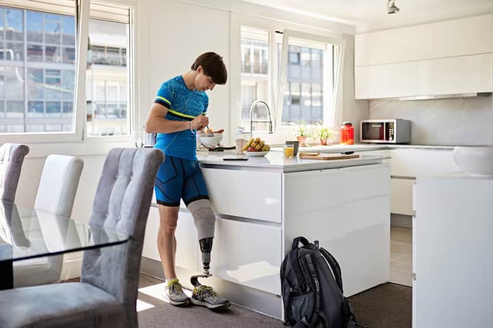 A person with a prosthetic leg eats breakfast in a modern kitchen. A backpack is on the floor nearby.