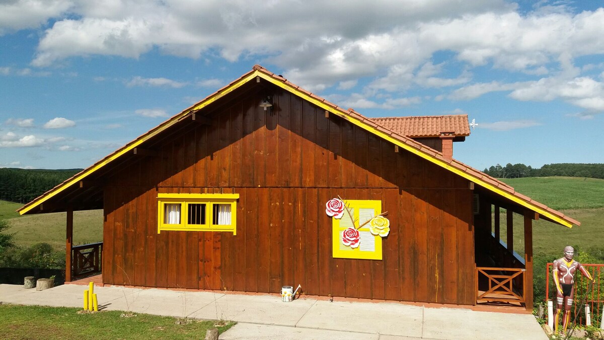 The exterior of a wooden house is displayed, featuring a warm brown color with yellow trim. Two charming flower designs adorn a window, enhancing the rustic charm. A covered porch offers a welcoming entry, while scenic fields stretch into the background under a partly cloudy sky.