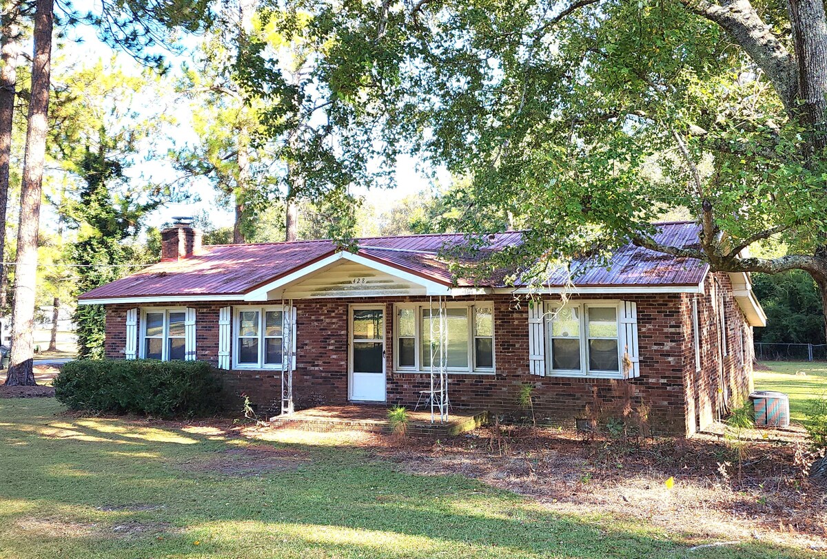 A brick bungalow is set in a landscaped yard, surrounded by tall trees. The house features multiple windows with white shutters, a front porch with swings, and a gabled roof. A grassy area extends around the home, creating a peaceful outdoor setting.