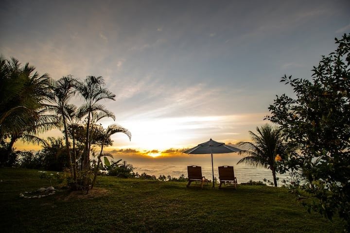 A serene outdoor setting is displayed with two lounge chairs positioned under a large umbrella. Lush greenery and palm trees surround the area, with a captivating view of the horizon where the sun rises over the ocean.