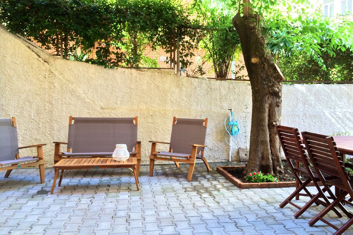 A private garden area features a wooden table surrounded by several folding chairs. Comfortable loungers are positioned nearby, set against a backdrop of lush greenery and a textured wall. The space is well-suited for outdoor relaxation and dining.