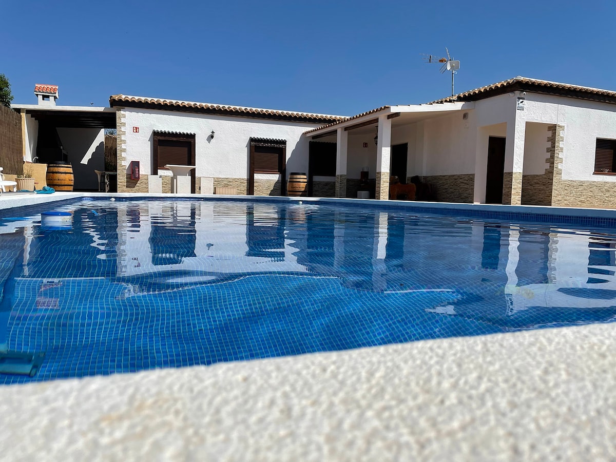 A large, clear swimming pool is seen in the foreground, featuring shimmering blue tiles. The background includes a single-story structure with multiple entrances and windows, surrounded by a sunlit outdoor space, emphasizing a peaceful and relaxing environment.
