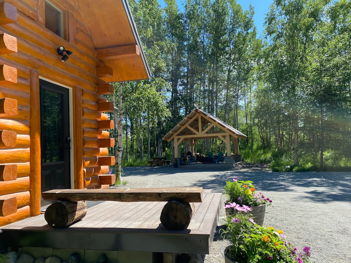 A modern log cabin is framed by lush greenery, showcasing a wooden deck with a sturdy log bench. In the background, a covered outdoor seating area is presented, surrounded by trees and gravel, creating a serene and inviting environment.
