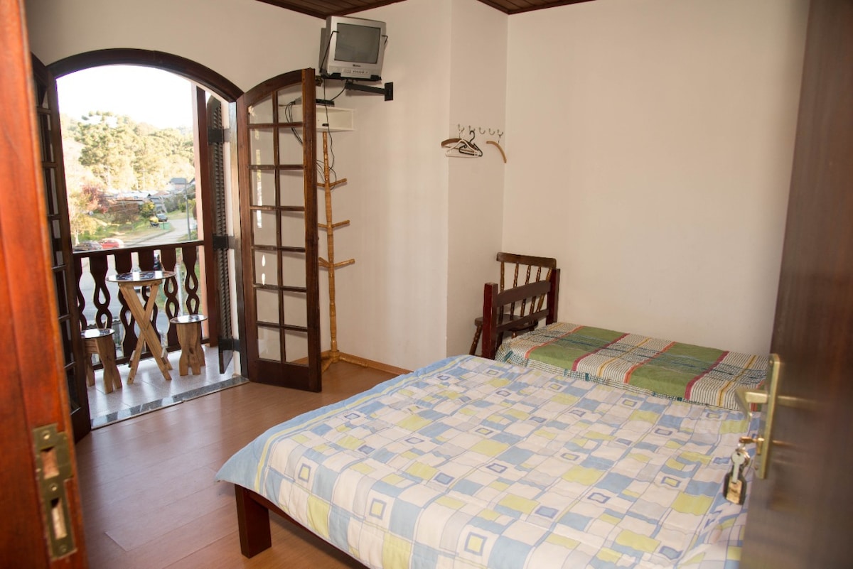 A simple bedroom features a spacious bed covered with a patterned quilt. A wooden chair and rack for hanging items are visible. Tall glass doors open to a small balcony, allowing natural light to fill the room.