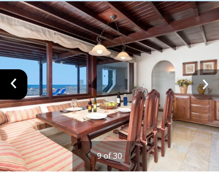 An inviting dining area features a wooden table surrounded by six high-backed chairs. Natural light enters through large windows, offering views of the sea. Adjacent, a wooden sideboard displays decorative items and flowers, enhancing the room's warm ambiance.