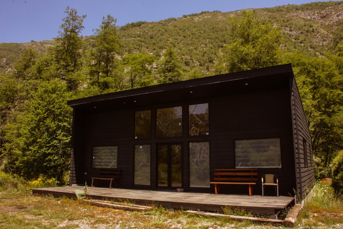 A wooden cabin is presented with large glass windows oriented towards the sun and mountains. The structure features a spacious deck area with minimal seating, surrounded by lush greenery and rolling hills, creating a serene outdoor setting.