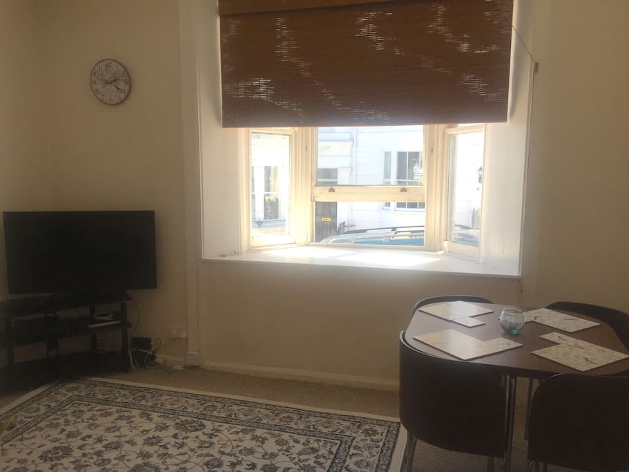 Living room photo showing dining table and bay window, to the right as you come through the door there is also a large standing mirror and a second fridge.