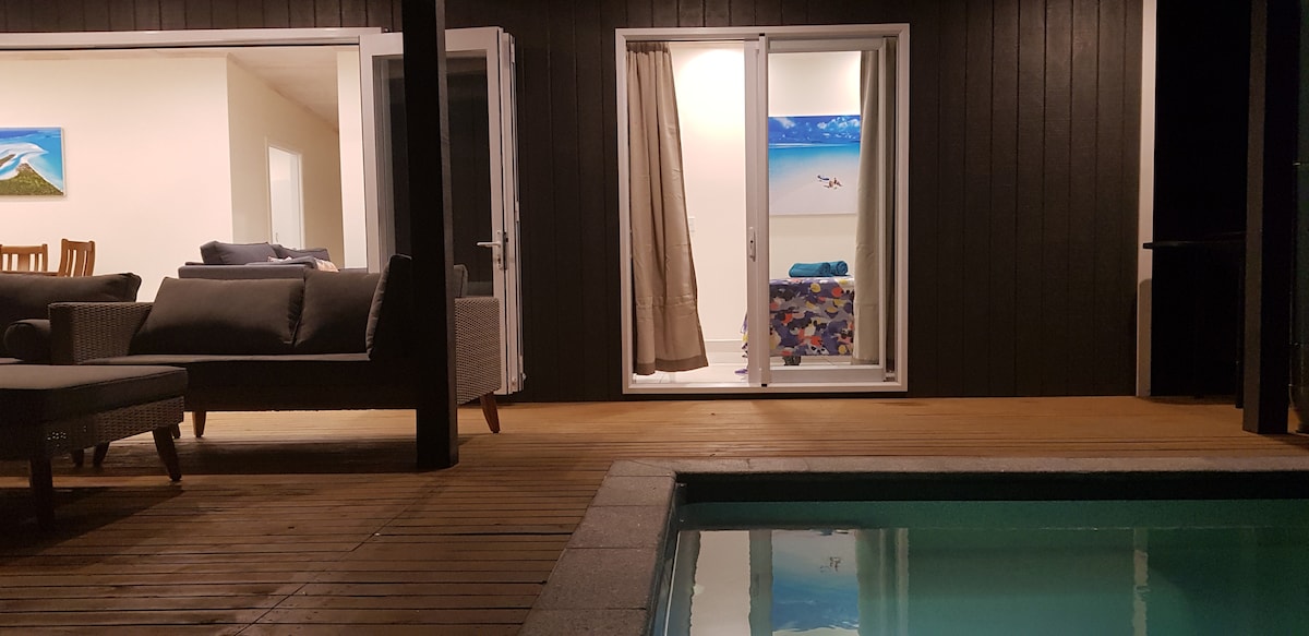 A view of the villa's outdoor area showcases a well-maintained wooden deck and a clear swimming pool. Large glass doors open to a cozy seating area, with a soft couch visible. Light-colored walls contrast with the dark wood exterior, and colorful artwork complements the serene ambiance.