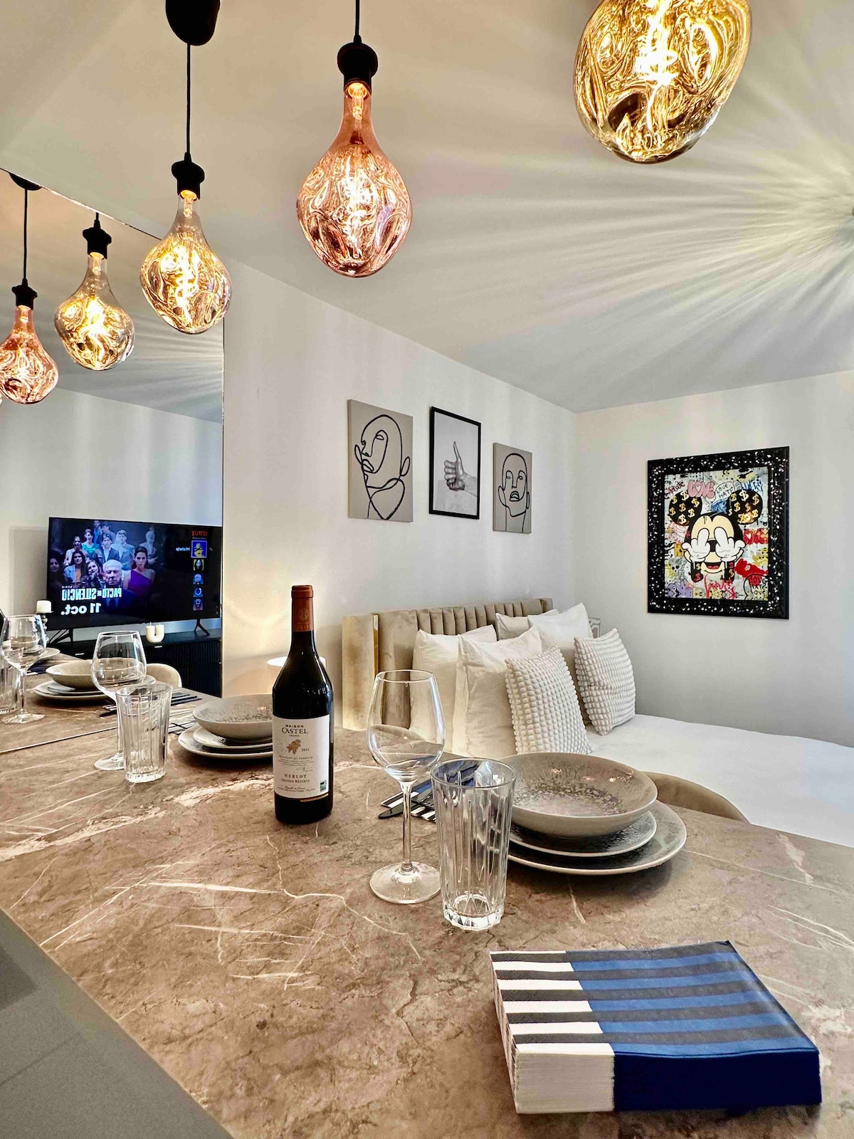 A modern dining area features a marble countertop with an inviting arrangement of dinnerware including plates and glassware. Pendant light fixtures with unique designs illuminate the space. A wall-mounted television is positioned nearby, while framed artworks add visual interest to the clean, neutral decor.
