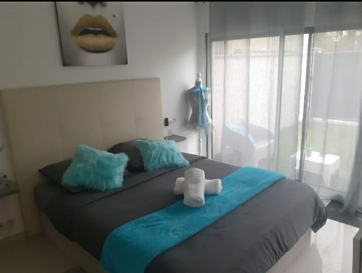 A spacious bedroom with a queen-size bed is presented, featuring soft grey linens accented by a vibrant turquoise throw. Decorative pillows enhance the bed's comfort. A large window allows natural light to fill the room, with a private outdoor space visible outside.