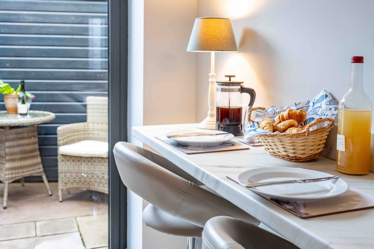 A dining area is shown with a smooth countertop and two sleek bar stools. A light source from a lamp softly illuminates a coffee press, a bottle of juice, and a basket filled with pastries. Outdoor seating is visible through the glass doors.