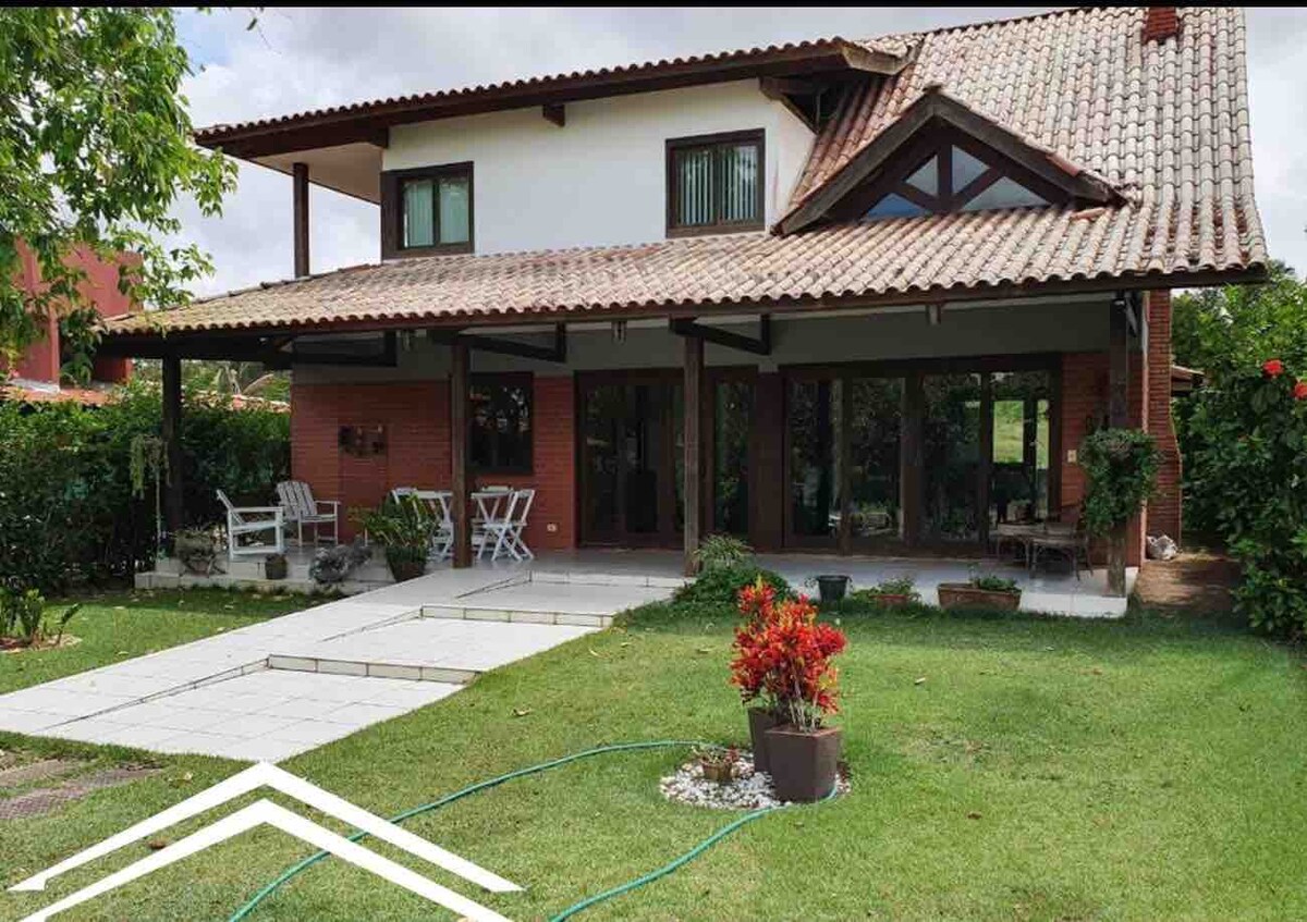 A spacious two-story house is surrounded by well-maintained greenery. The entrance features a stone pathway leading to a covered patio with outdoor seating. The exterior showcases a tiled roof and large glass windows, creating a bright and open atmosphere.