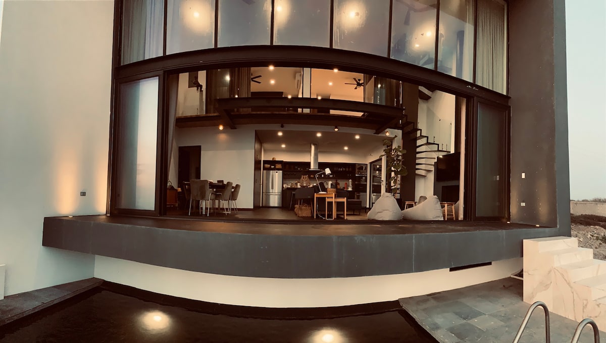 A modern living space features large glass sliding doors that open to a serene outdoor area, enhancing the connection to nature. The interior showcases a dining table, comfortable seating, and a stylish kitchen. Soft ambient lighting creates an inviting atmosphere throughout.