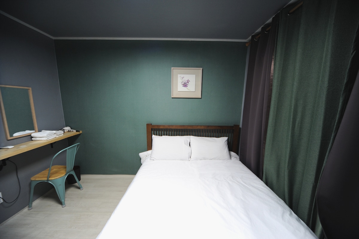 A simple sleeping area features a spacious bed dressed in crisp white linens. A green accent wall complements the dark curtains, while a small desk with a mirror and a chair is positioned nearby, contributing to a functional and comfortable environment.