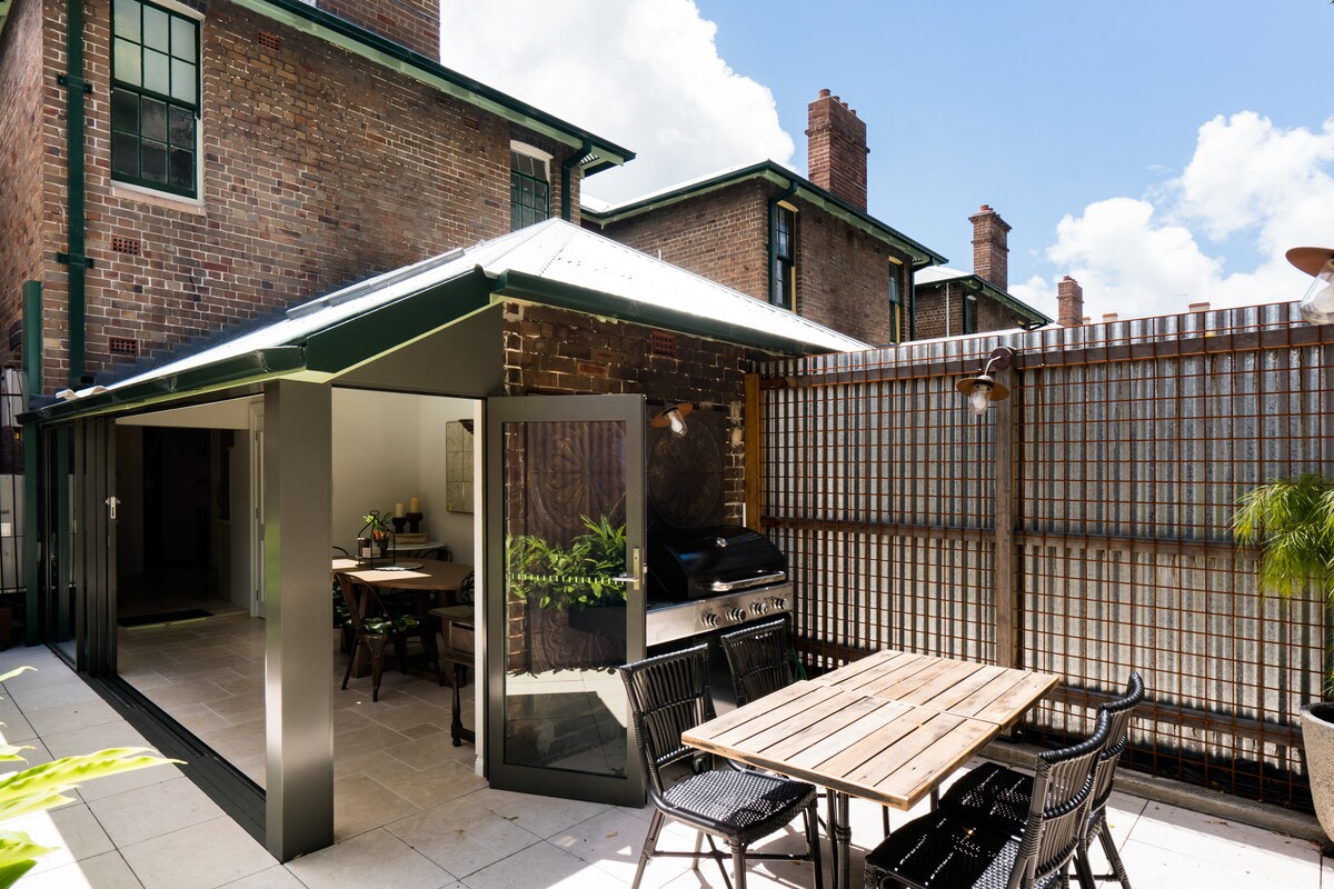 An outdoor courtyard features a wooden dining table surrounded by black chairs. A Beefeater BBQ is positioned nearby, with a glass sliding door leading into an interior space. The surrounding walls are designed with a lattice pattern, complementing the brick structure.