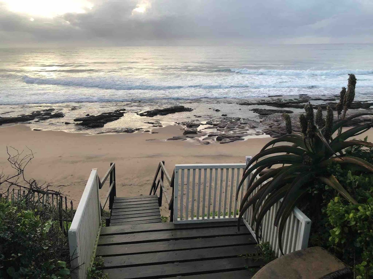 A wooden staircase leads down to the beach, flanked by lush greenery. The sandy shore is visible, with gently rolling waves lapping at the water's edge. Rocky formations are present, and the sky is filled with soft, cloudy hues reflecting the early morning light.