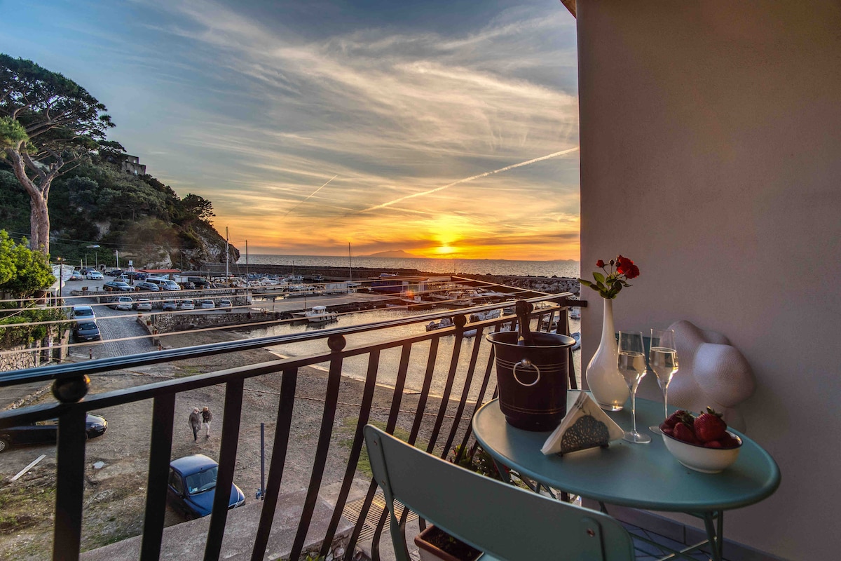 A small balcony features a round table set for two, complete with glasses and strawberries. The view overlooks a tranquil harbor with boats and the sea, framed by a sunset sky filled with warm hues. Greenery and a coastal road are visible in the background.