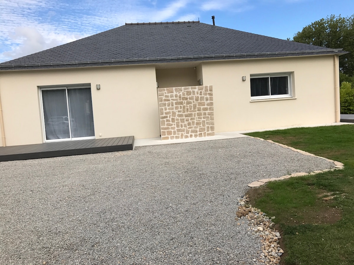 The exterior features a modern single-story home with a sloped roof. A paved area is visible in front, surrounded by a gravel driveway. A small stone wall provides privacy near the entrance, while a neatly maintained lawn offers a green contrast to the gravel.