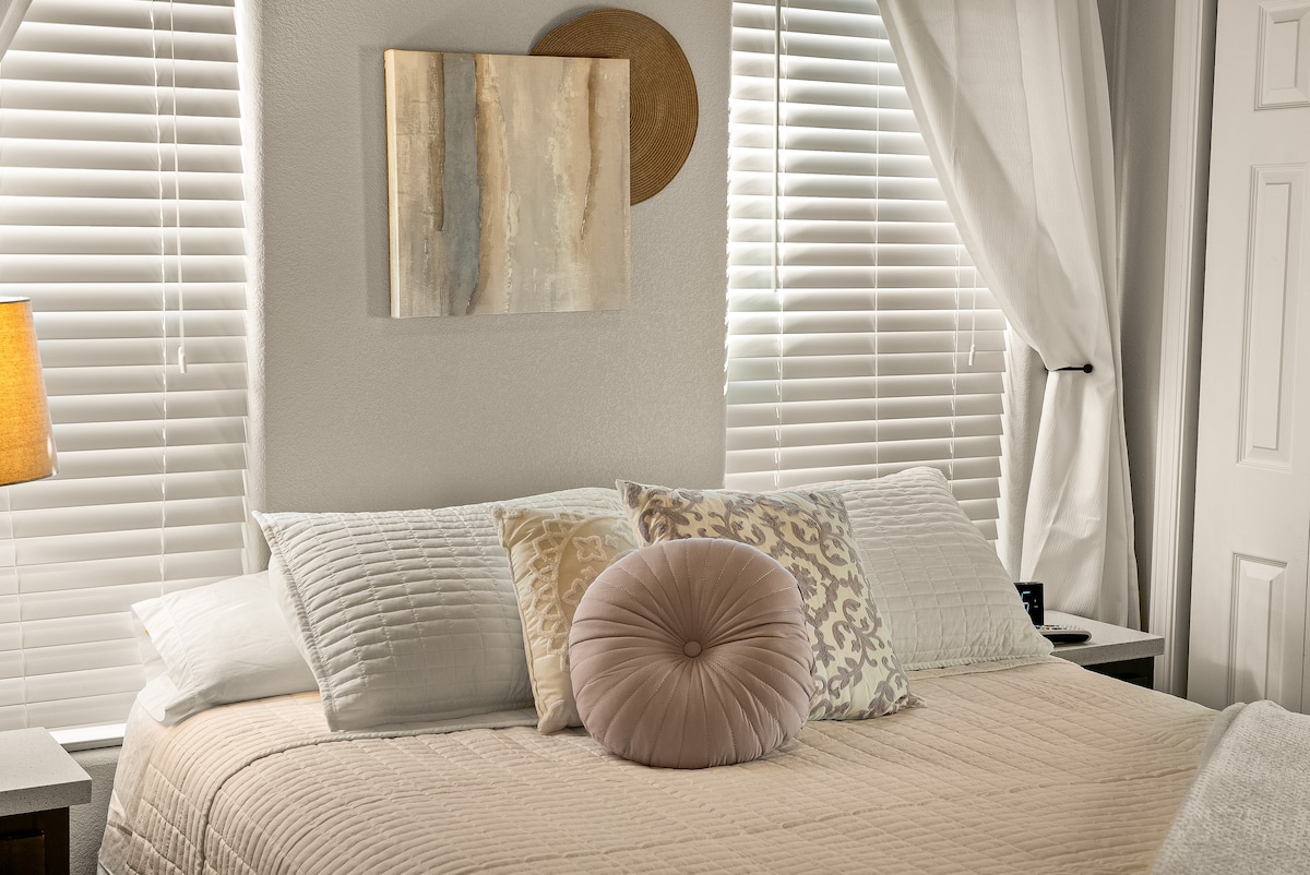 A well-appointed sleeping area features a spacious bed topped with decorative pillows in soft hues. Natural light filters through partially closed blinds, which frame the room. A minimalist artwork and wall decor, including a woven plate, add subtle elegance.