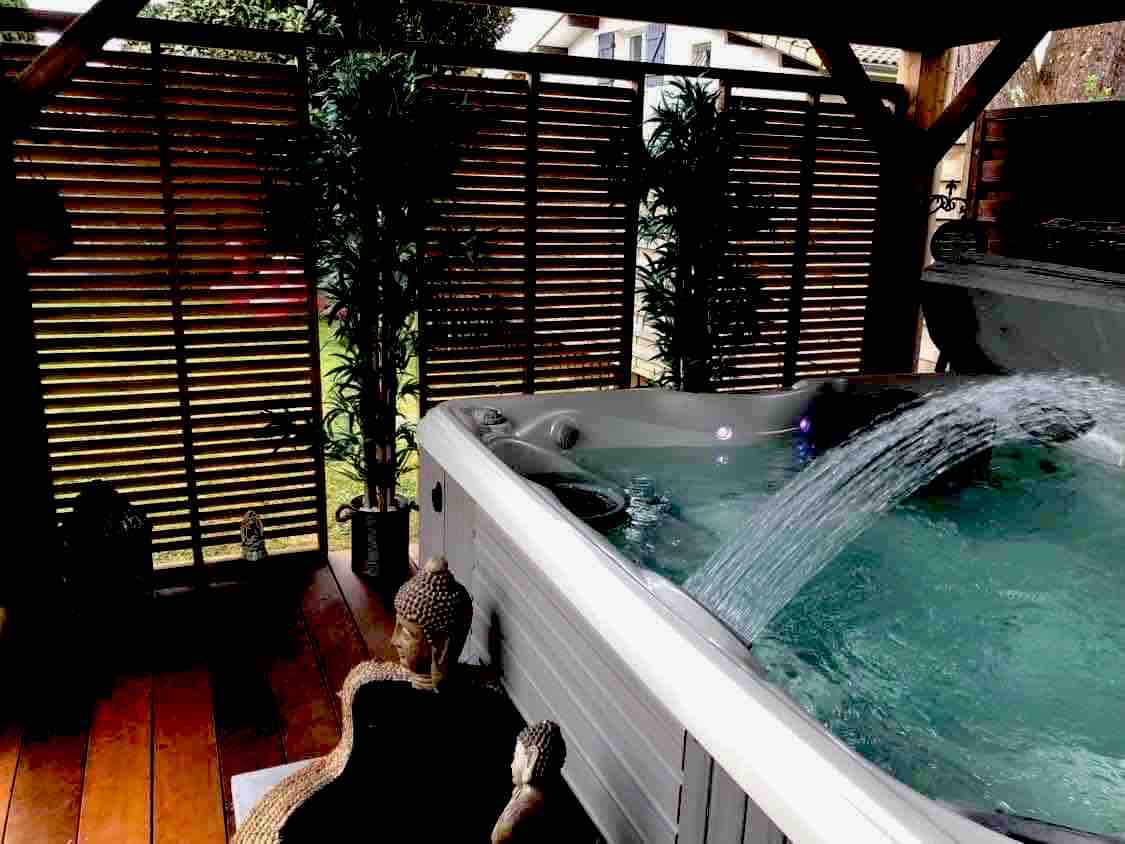 An outdoor jacuzzi is visible, featuring bubbling water and gentle jets. Surrounding greenery adds a touch of nature, while wooden slats create privacy. The deck is adorned with potted plants, contributing to a tranquil environment ideal for relaxation.