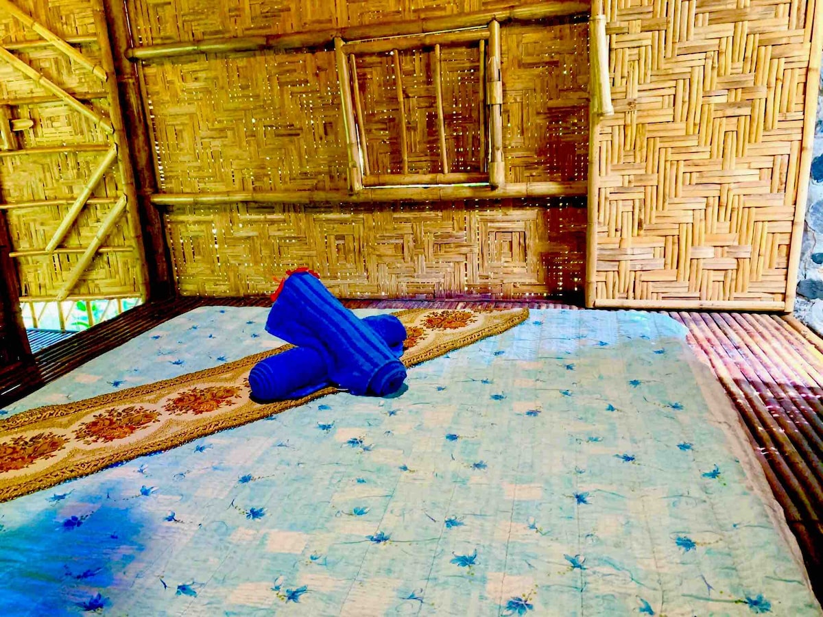 A bamboo interior is showcased, featuring a spacious bed covered with a light blue patterned blanket, complemented by rolled towels. A woven bamboo wall provides a natural aesthetic, while soft lighting creates a calming environment.