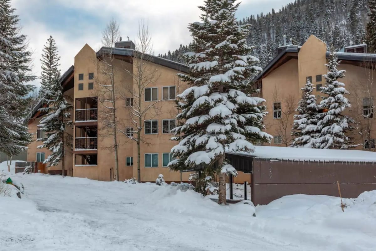 A spacious three-story condo building is surrounded by snow-covered trees, creating a serene winter scene. The structure features multiple windows, with balconies visible on the upper floors. The entrance is approached via a snowy path, adding to the inviting atmosphere of the area.