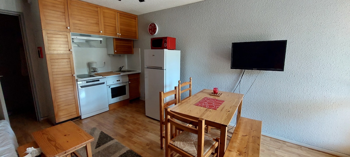 The living area features a functional kitchen with wooden cabinets, a refrigerator, and a microwave. A dining table is set for four, complemented by a television mounted on the wall. Wooden floors contribute to a warm and cohesive atmosphere throughout the space.