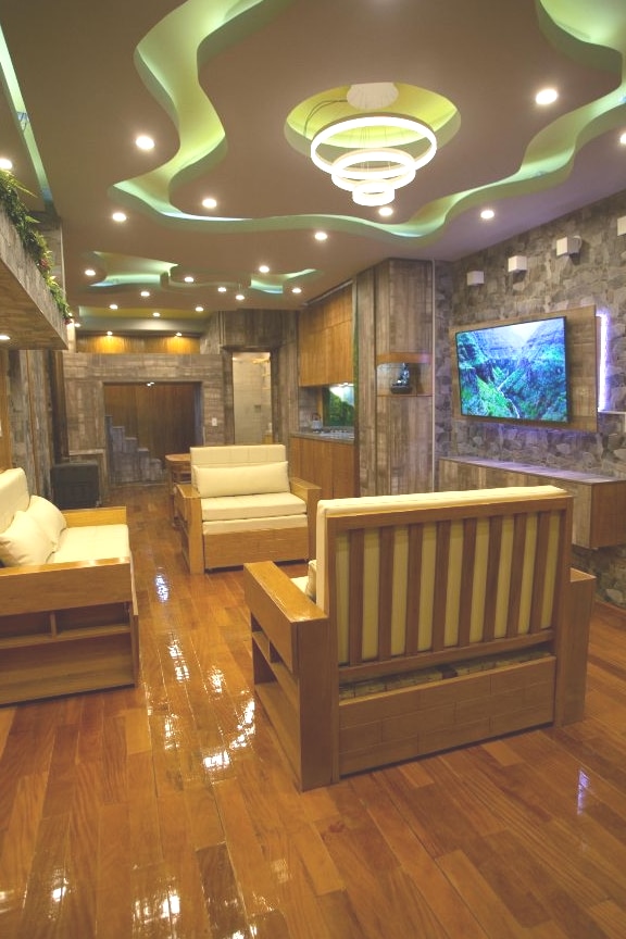 A modern living area is featured, showcasing wooden seating arrangements with neutral cushions. A stylish chandelier illuminates the space, complemented by green ceiling accents. A large wall-mounted TV displays scenic imagery, while natural stone walls create a warm and inviting backdrop.