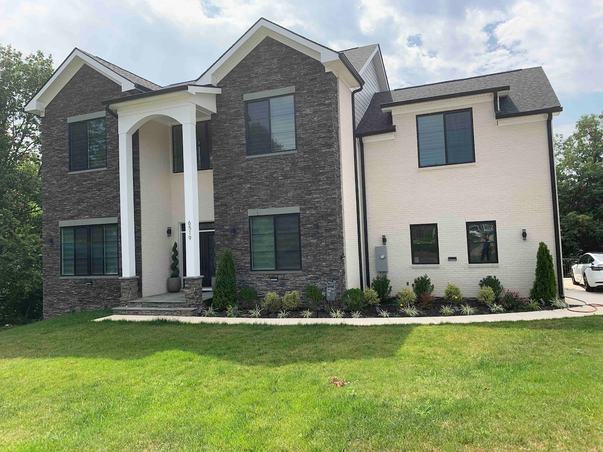 The exterior of a modern two-story home is presented, showcasing a blend of dark stone and white siding. Large windows reflect natural light, and a neatly landscaped yard features green grass and a flower bed. The entrance is marked by a covered porch.