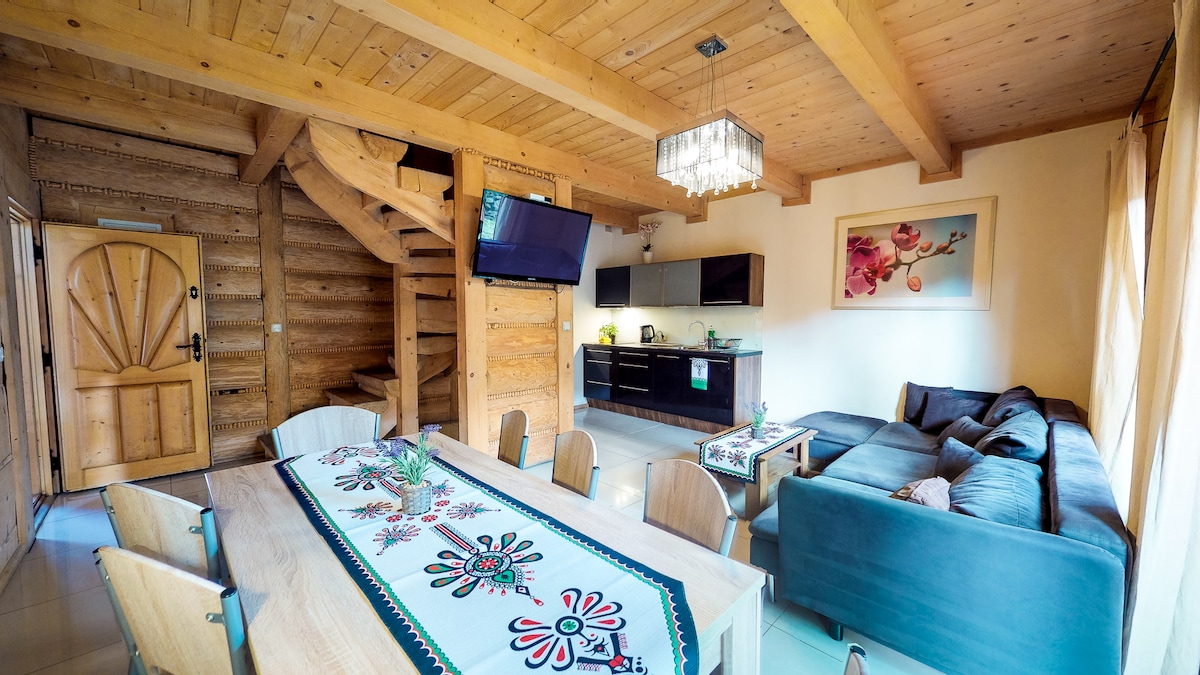 An open-concept living space showcases wooden beams and a rustic staircase. A long dining table is set with decorative elements, while a comfortable sofa rests nearby. Large windows allow natural light to fill the room, enhancing the contemporary kitchen equipped with modern appliances.