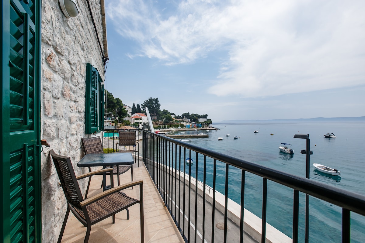 A balcony overlooking the sea features a table with two chairs, framed by decorative green shutters. The view includes boats floating on the calm water and the scenic shoreline, adding to the tranquil atmosphere of the coastal setting.