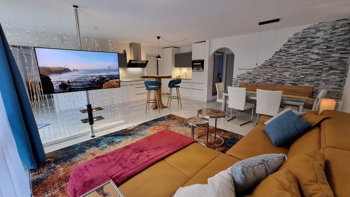 An open living area features a spacious sofa with a vibrant throw, facing a large wall-mounted television. The kitchen is visible in the background, complemented by bar stools at a stylish counter. A dining table with modern chairs is situated near the patterned rug.