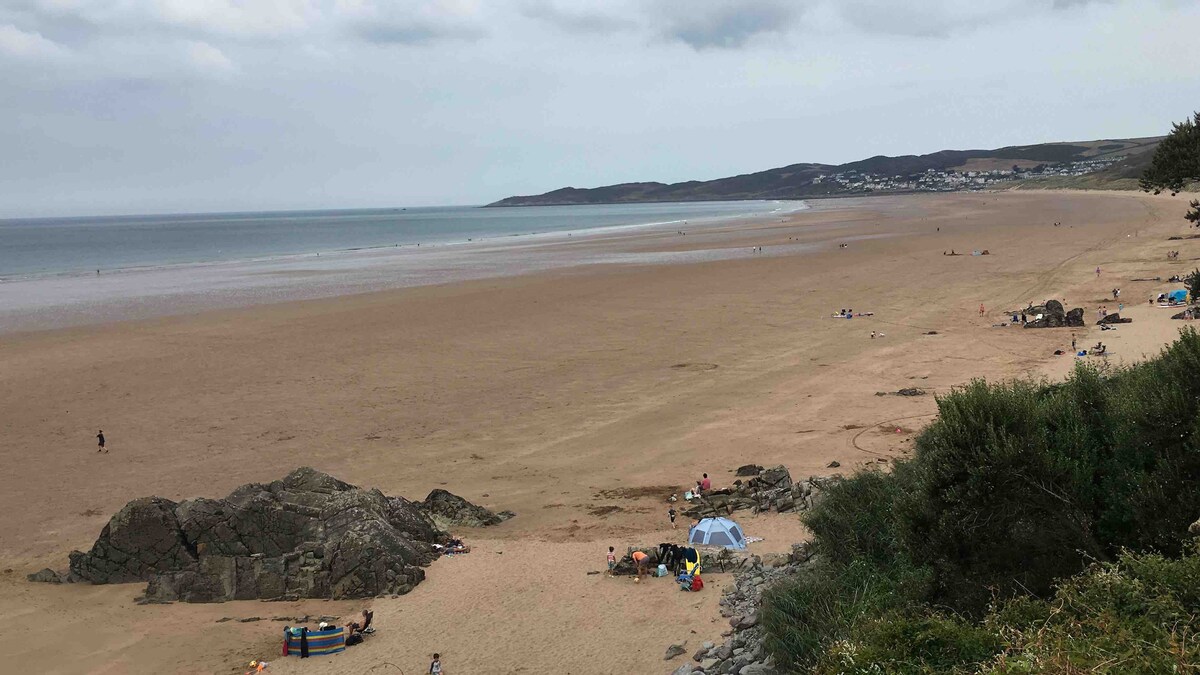 A vast sandy beach stretches along the coastline, with gentle waves lapping at the shore. A few small groups of people are scattered across the beach, alongside colorful umbrellas and beach chairs. The distant hills and village are visible under a cloudy sky, creating a serene atmosphere.