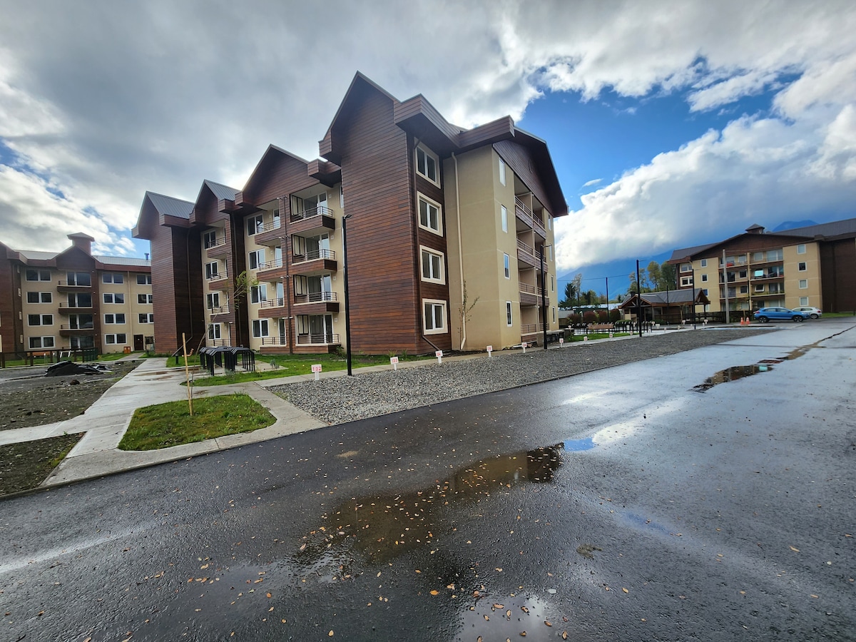 A modern apartment building is depicted with a mix of wood and light-colored facades. The exterior features multiple balconies and large windows. A rain-slicked driveway and landscaped areas are visible in the foreground, with a cloudy sky providing a backdrop.