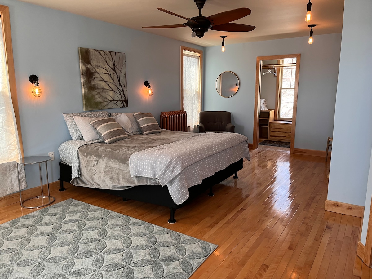 A spacious bedroom features a king bed with light grey bedding and decorative pillows. Natural wood floors and large windows enhance the bright atmosphere. A comfortable chair, round mirror, and a modern lamp complete the room, providing ample space for relaxation.