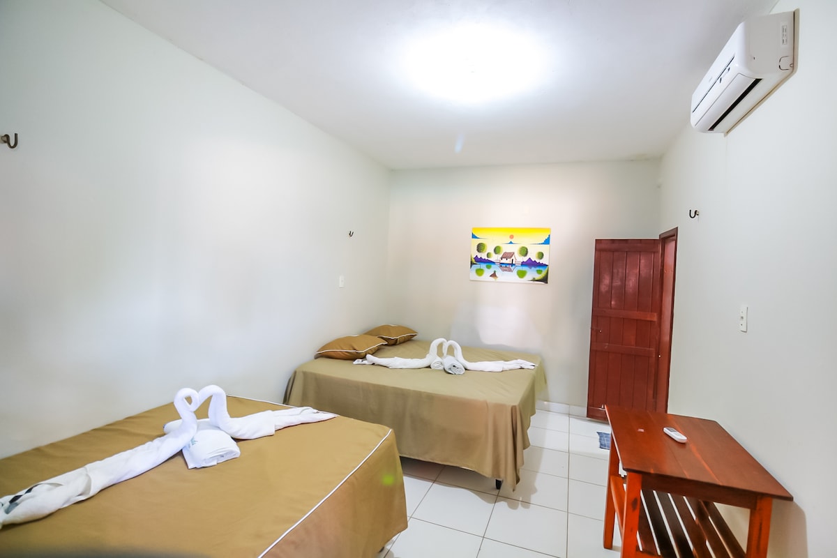 A simple and inviting guest room features two spacious beds dressed in neutral-colored linens. A vibrant piece of wall art adds a pop of color on one side, while a wooden shelf is positioned nearby. Air conditioning is present for comfort.
