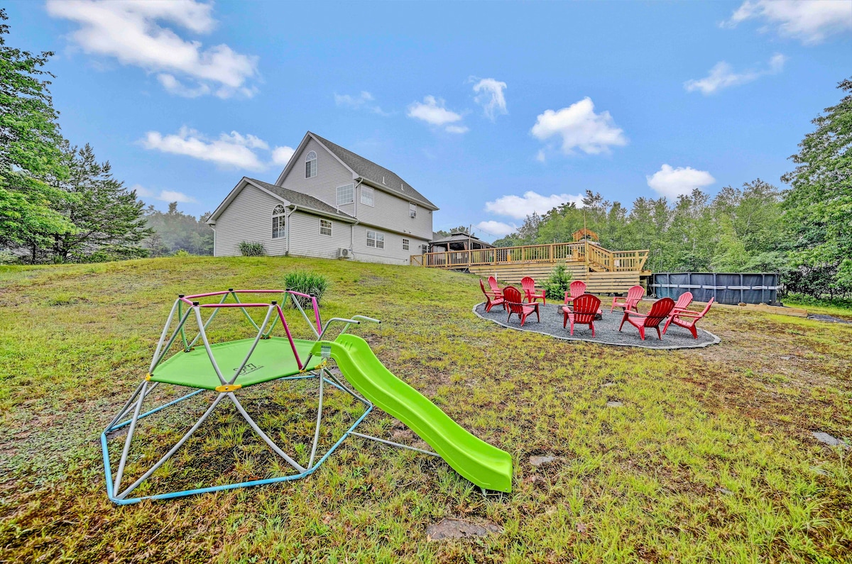 An outdoor area features a green slide and a climbing structure, accompanied by a circle of six red Adirondack chairs arranged around a fire pit. The spacious backyard is surrounded by trees, offering a serene setting with a deck visible in the background.