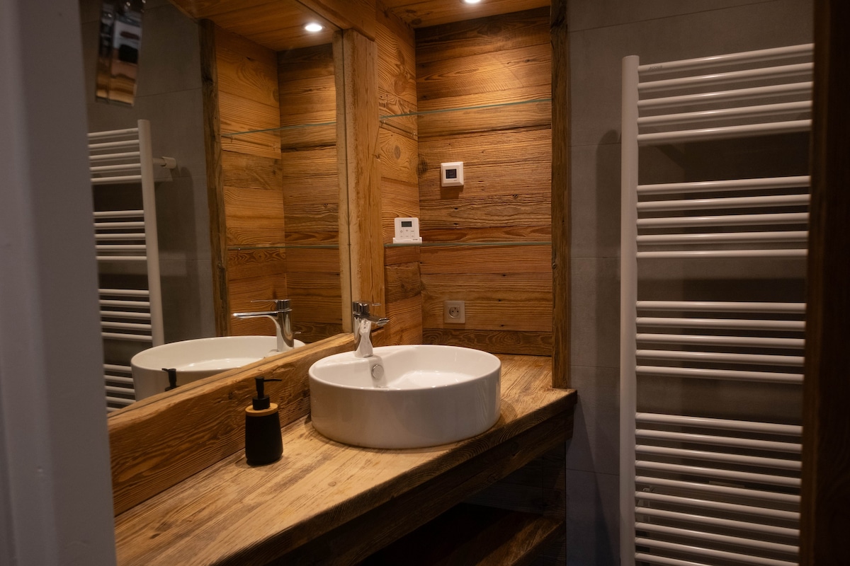 A modern bathroom features wooden paneling and warm lighting. A circular sink is set atop a wooden countertop, with a wall mirror providing additional light. A sleek towel radiator is mounted to the wall, complementing the natural materials.