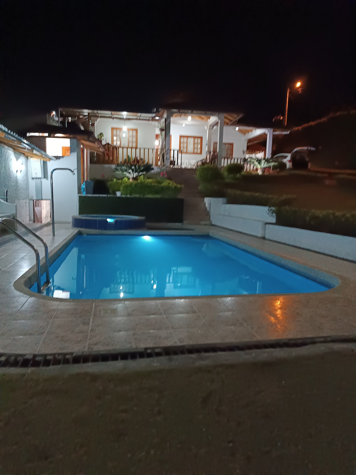 A tranquil swimming pool is illuminated at night, reflecting soft blue light. Surrounded by tiled flooring, the pool is set against a backdrop of a welcoming house with multiple windows and a landscaped area featuring greenery.