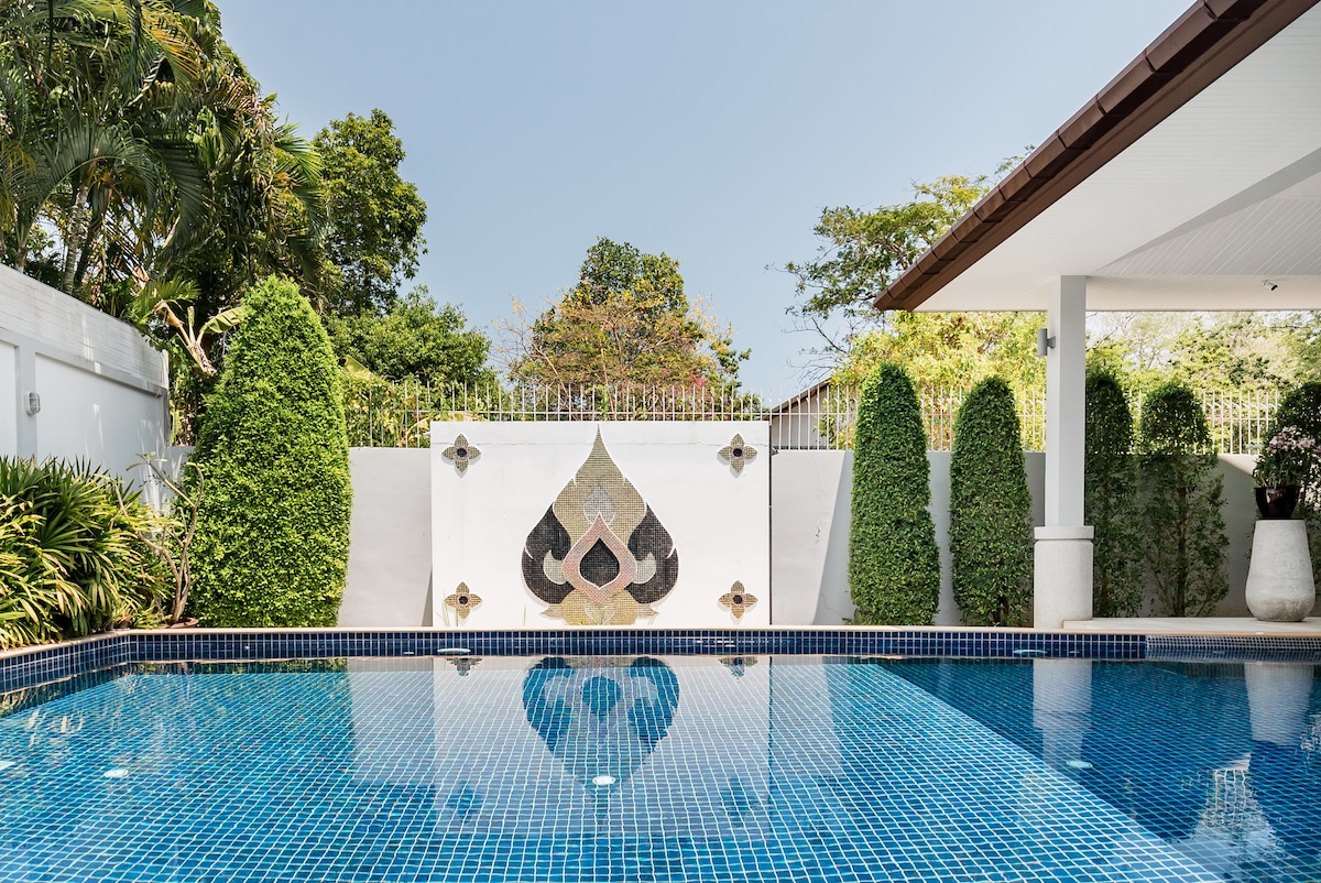 A sparkling lap pool reflects the clear sky, framed by lush greenery and well-manicured hedges. A decorative wall with intricate patterns adds a touch of artistry, enhancing the serene outdoor environment.