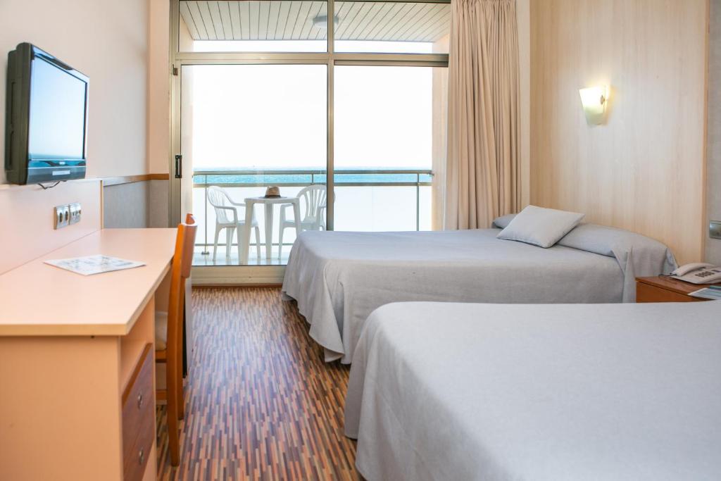 A spacious guest room features two double beds with soft gray bedding. A small balcony is visible through large glass doors, offering a seating area with a table and chairs. A television and a wooden dresser are along one wall, complemented by a warm wood floor.