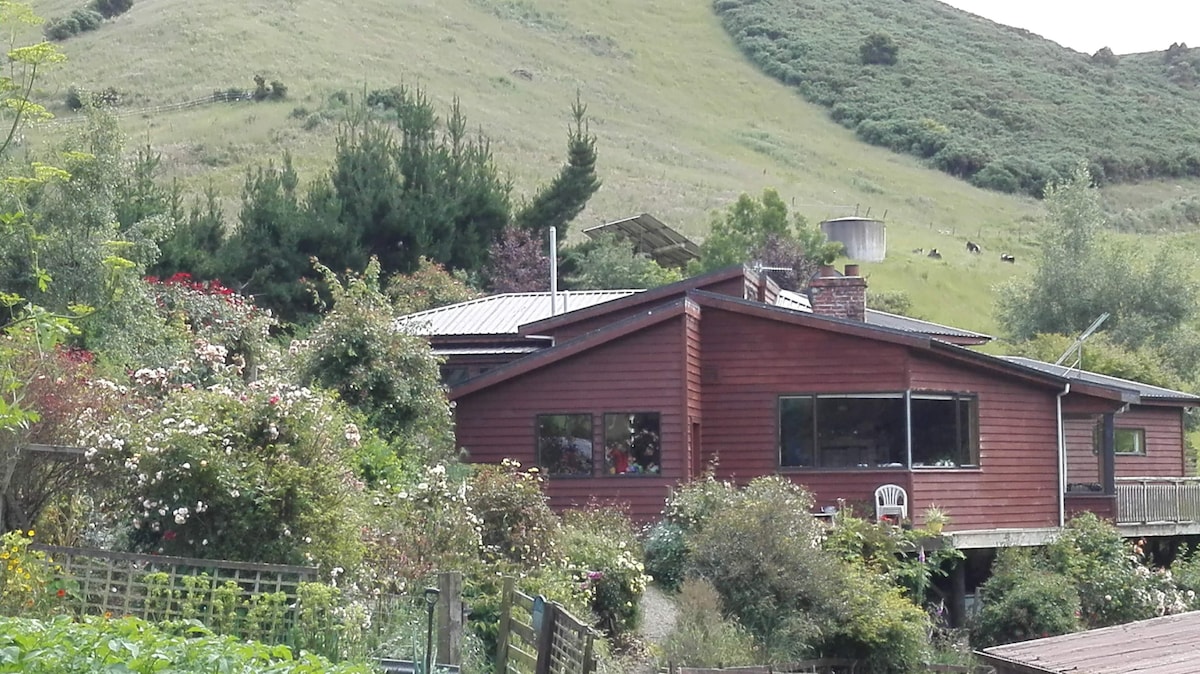A wooden farmhouse is nestled against a gently sloping hillside, surrounded by lush greenery and colorful flowers. Large windows offer views of the surrounding landscape. A deck extends from the house, providing an outdoor relaxation area with views of the farm and hills beyond.