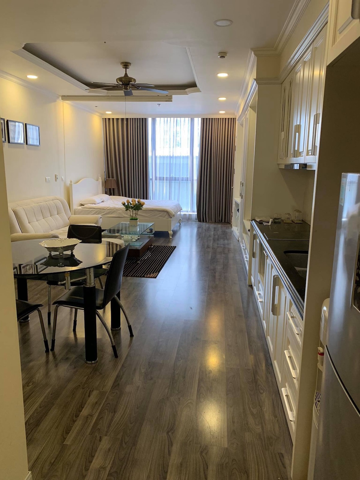A bright and modern studio is presented, featuring a spacious living area with a comfortable sofa beside a dining table and chairs. The kitchenette is visible along one side, with white cabinetry offering ample storage. Large windows allow natural light to fill the space.