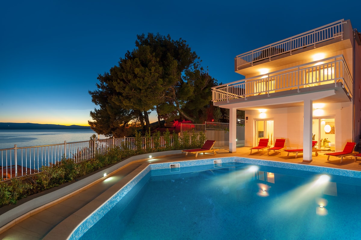 A serene outdoor area is highlighted, featuring a spacious pool with illuminated water and comfortable lounge chairs. The villa's elegant structure and balconies showcase a stunning sea view against a twilight sky, surrounded by lush greenery and gentle slopes.