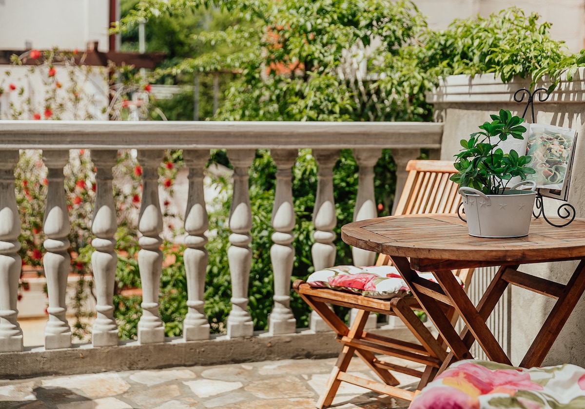 A terrace area is depicted, featuring a wooden table with a small potted plant at its center. Two matching wooden chairs, adorned with floral cushions, complement the setting. Lush greenery frames the space, enhancing the connection to the garden.