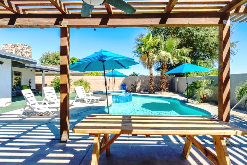 An outdoor area features a spacious swimming pool surrounded by palm trees and lounge chairs. A shaded picnic table offers seating under wooden beams, complemented by vibrant blue umbrellas providing shade. The inviting pool reflects sunlight, creating a serene atmosphere for relaxation.