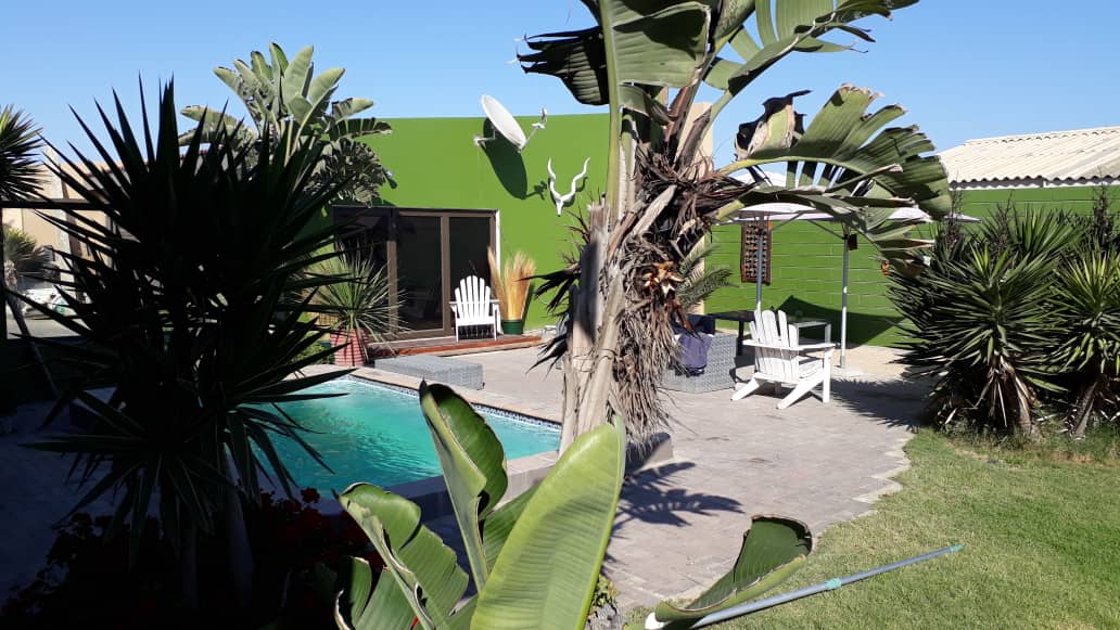 A vibrant green structure is framed by tropical vegetation, featuring a private pool surrounded by lounging chairs. Sunlight casts a warm glow over the outdoor area, enhancing the serene environment. A mix of palms and other greenery fills the space, creating a relaxing retreat.