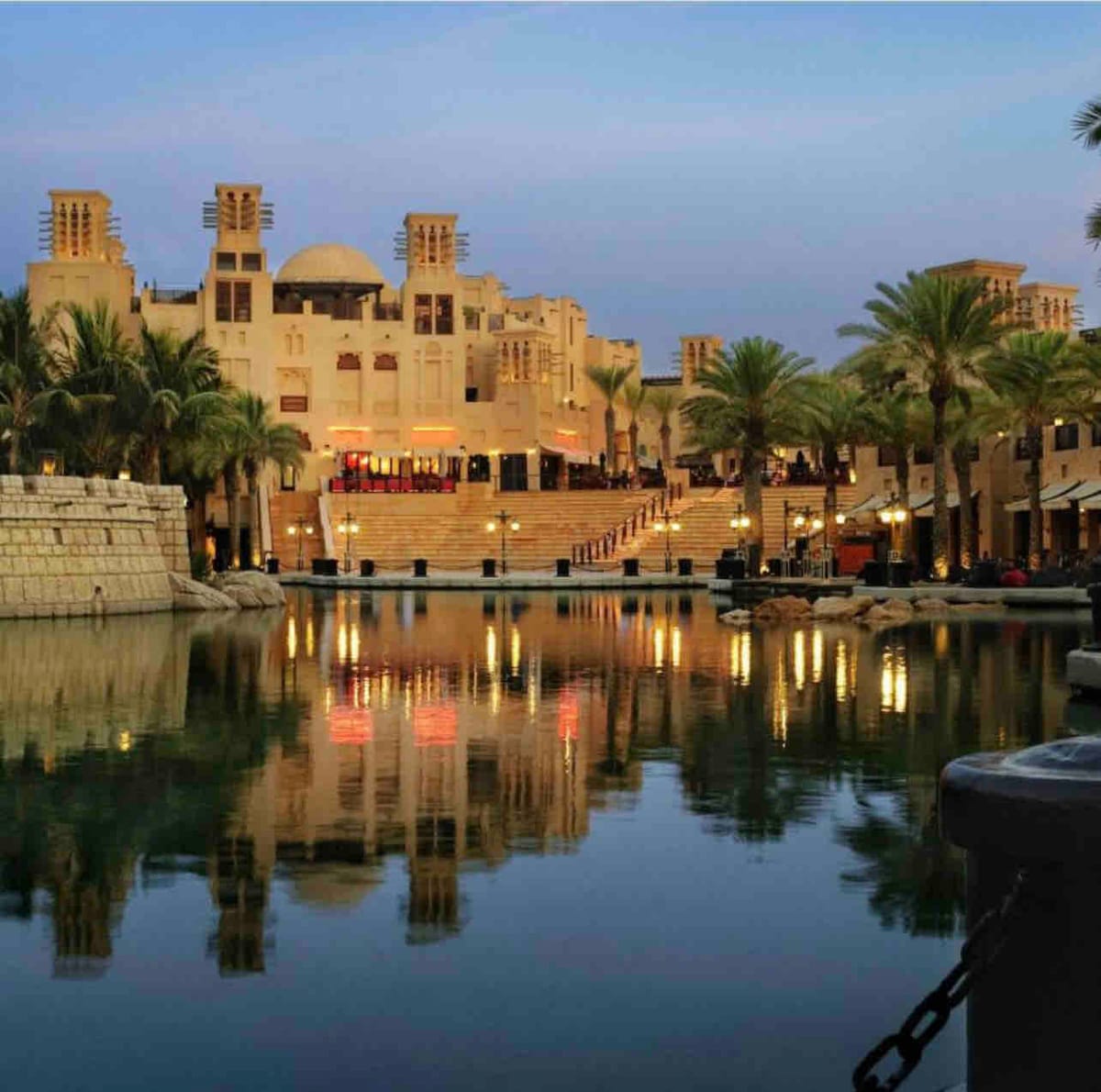 The image captures an elegant waterfront scene at dusk, showcasing modern architectural elements reflecting on calm waters. Lush palm trees frame the area, while warm lights illuminate the steps leading to terraced dining spaces. The serene atmosphere is enhanced by the surrounding tranquil landscape.
